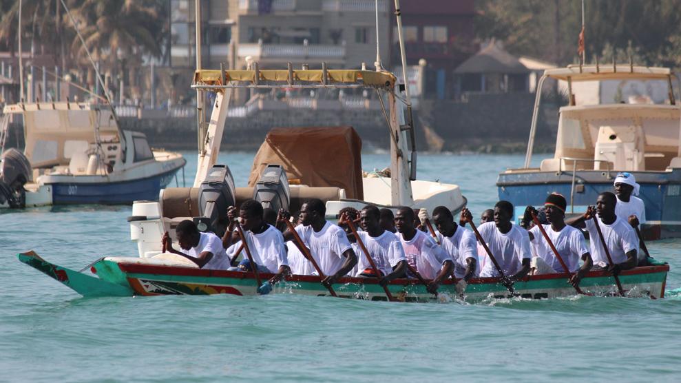 A team of rowers in white shirts and black caps paddling in unison in a long boat, with other boats and buildings in the background.